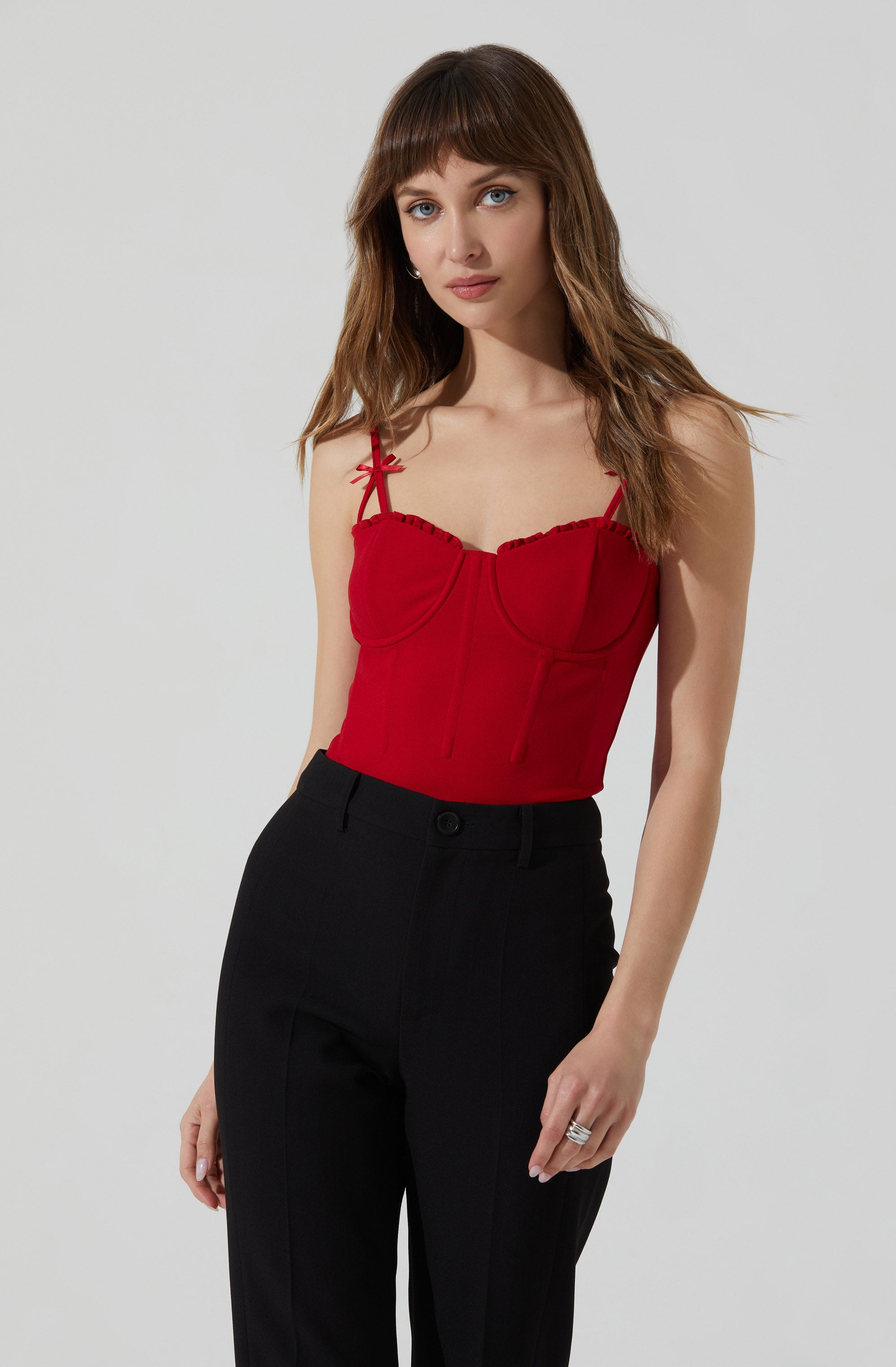 A woman is wearing a red sleeveless top with thin, tied shoulder straps and a structured, bustier-style design. She pairs it with high-waisted black trousers.