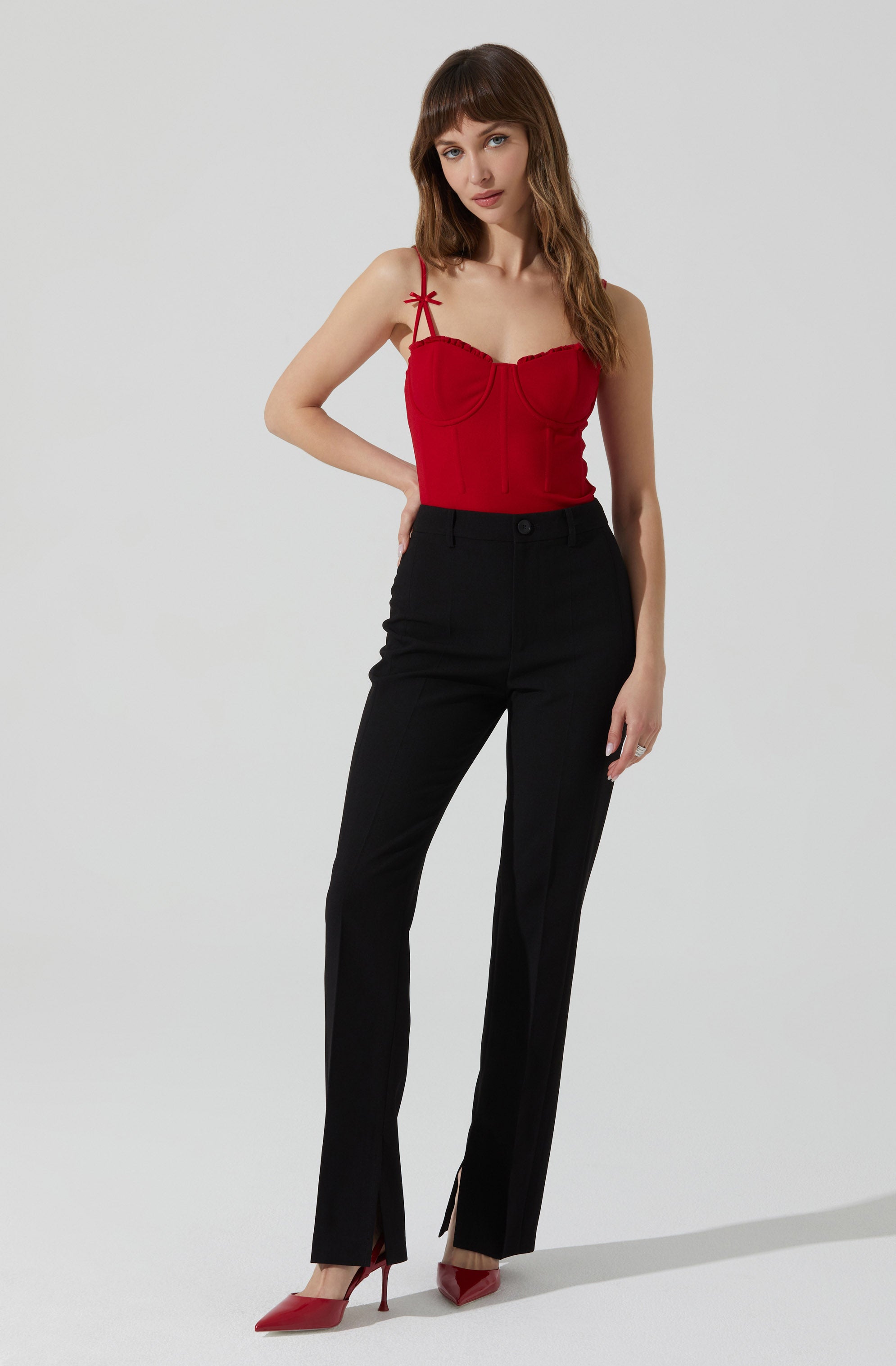 A woman is wearing a red sleeveless top with thin straps and small bows on the shoulders. She pairs it with black high-waisted trousers that have a straight leg fit and a slit at the bottom. She is also wearing red pointed-toe high heels.