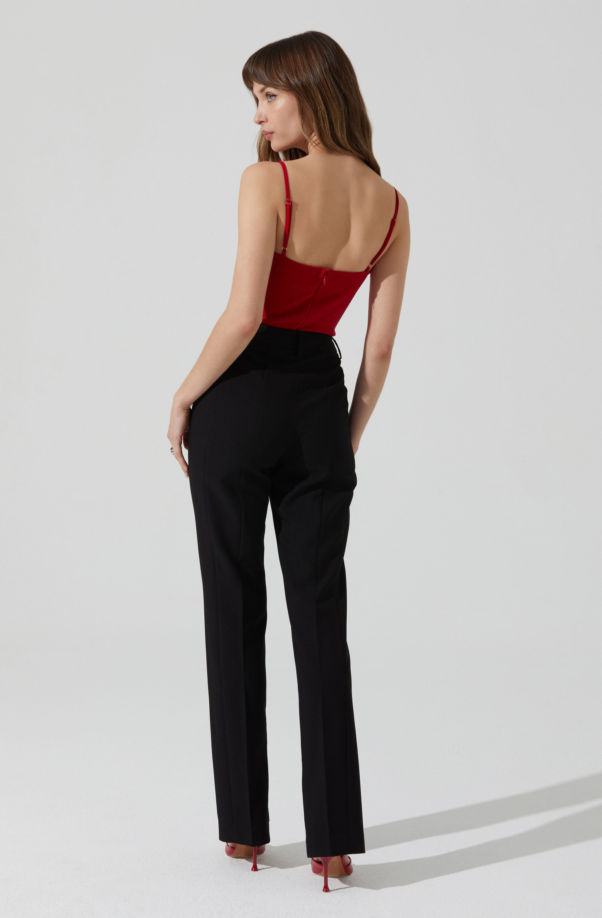 A woman is wearing a fitted red spaghetti strap top and high-waisted black tailored pants. She has long brown hair with bangs and is standing against a plain light background, with her back slightly turned to the camera. The red top features thin straps and a zipper at the back. The black pants have a classic straight-leg cut. She is also wearing red high-heeled shoes.