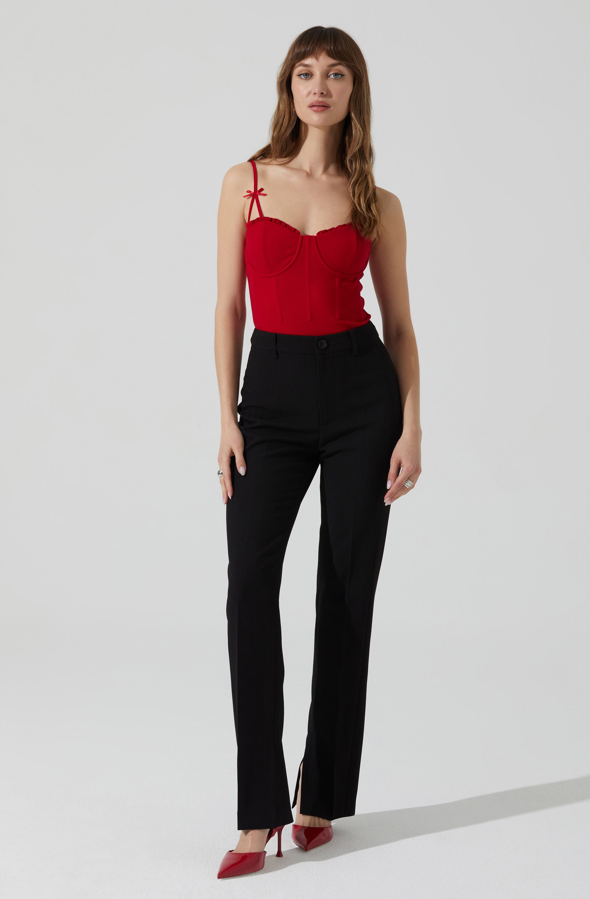 A woman is wearing a red sleeveless top with thin spaghetti straps, which are tied into small bows on her shoulders. The top has a fitted, bustier-style design with decorative seams. She pairs it with high-waisted black trousers that have a straight-leg cut and a slight slit at the ankle. She is also wearing red high-heeled shoes.