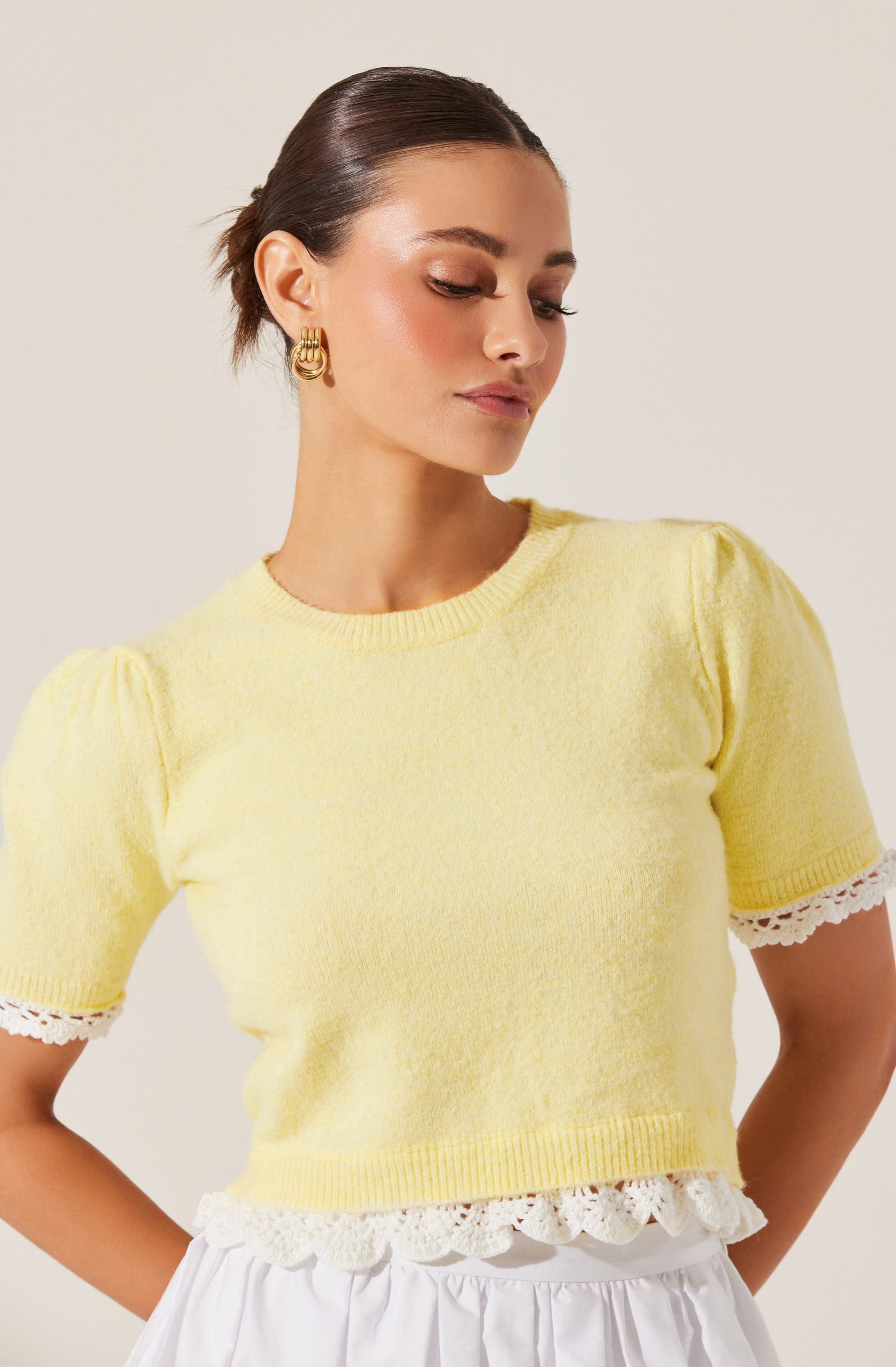 A woman is wearing a light yellow, short-sleeved knit top with puffed shoulders. The top has a round neckline and features white lace trim with a scalloped edge at the hem and the sleeve cuffs. She is also wearing large, gold hoop earrings.