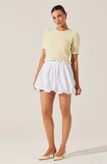 A woman is wearing a light yellow short-sleeved knit top with a round neckline and lace trim on the sleeve cuffs and hem. She is also wearing a white, pleated, knee-length skirt with a gathered waist. She has beige pointed-toe high heels.