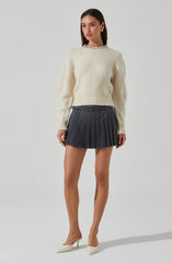 A young woman is wearing a fluffy, cream-colored, long-sleeve sweater with a decorative collar. She pairs it with a high-waisted, dark gray pleated mini skirt that has a waistband with belt loops and a button closure. She is also wearing light-colored high heel shoes.