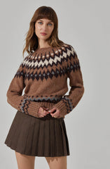 A woman is wearing a cozy, oversized sweater with a patterned design featuring white, black, and shades of brown across the chest and cuffs. The sweater is brown with a fair isle pattern around the yoke and sleeve cuffs. She pairs it with a dark brown plaid mini skirt with pleats.