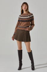 A woman is wearing a cozy, multicolored knit sweater with a pattern of diamonds and zigzags in shades of brown, beige, white, and dark blue. She pairs it with a high-waisted, pleated mini skirt in a dark brown houndstooth pattern. She completes the outfit with tall, black leather knee-high boots.