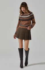 A woman is wearing a cozy, patterned knit sweater in shades of brown, cream, black, and gray, with a geometric design around the yoke and cuffs. She has paired it with a high-waisted, pleated mini skirt in a brown checkered pattern. She is also wearing tall black leather boots that reach just below the knees.