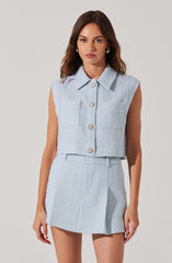 A young woman is wearing a light blue sleeveless cropped top with a collar and four decorative buttons down the front, paired with a matching high-waisted, A-line mini skirt. The outfit is made of a textured, woven fabric.