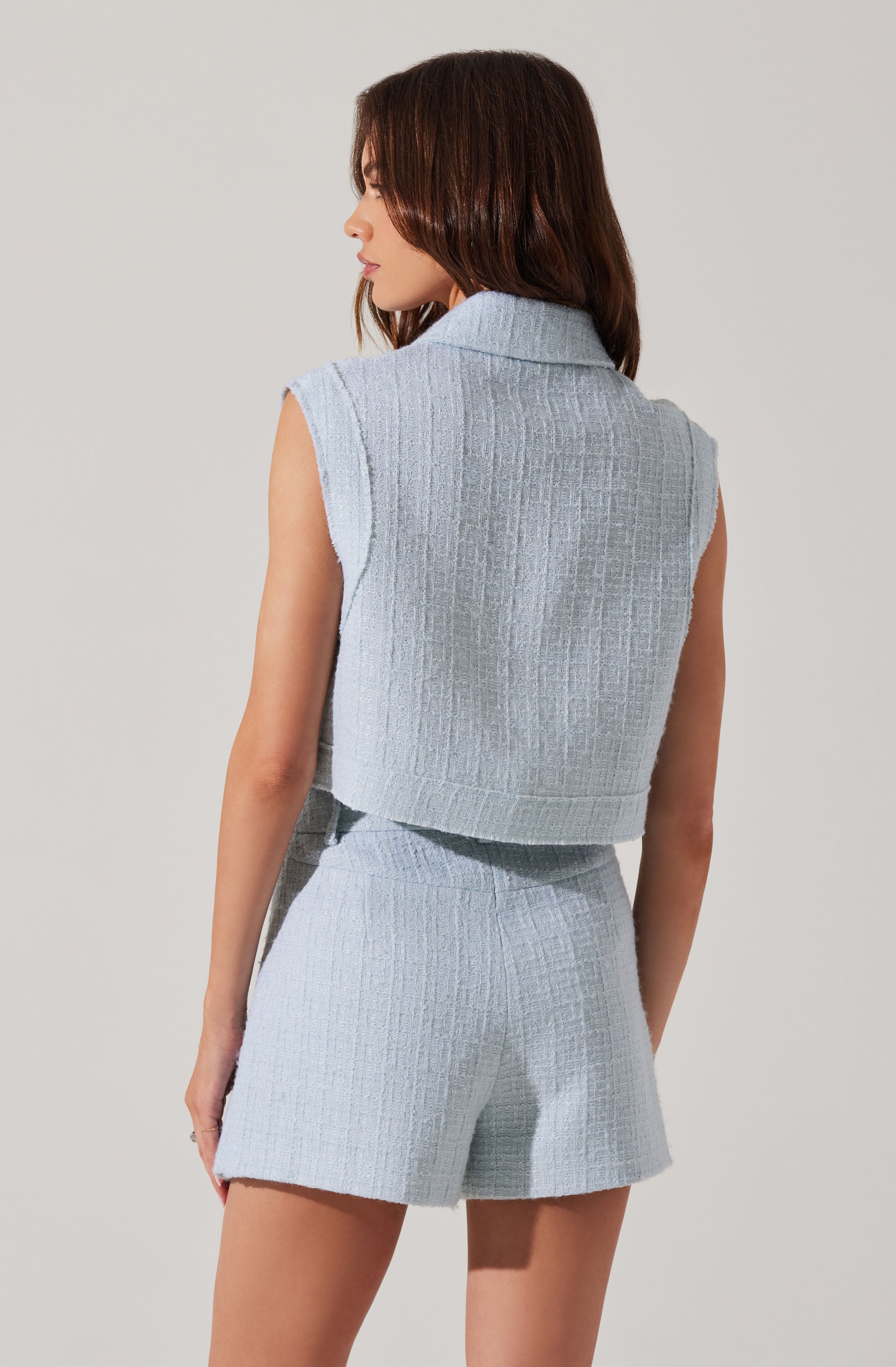 A woman is wearing a sleeveless, light blue textured knit vest with a collar, paired with matching high-waisted shorts that have the same textured knit pattern.
