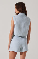 A woman is wearing a sleeveless, light blue textured knit vest with a collar, paired with matching high-waisted shorts that have the same textured knit pattern.