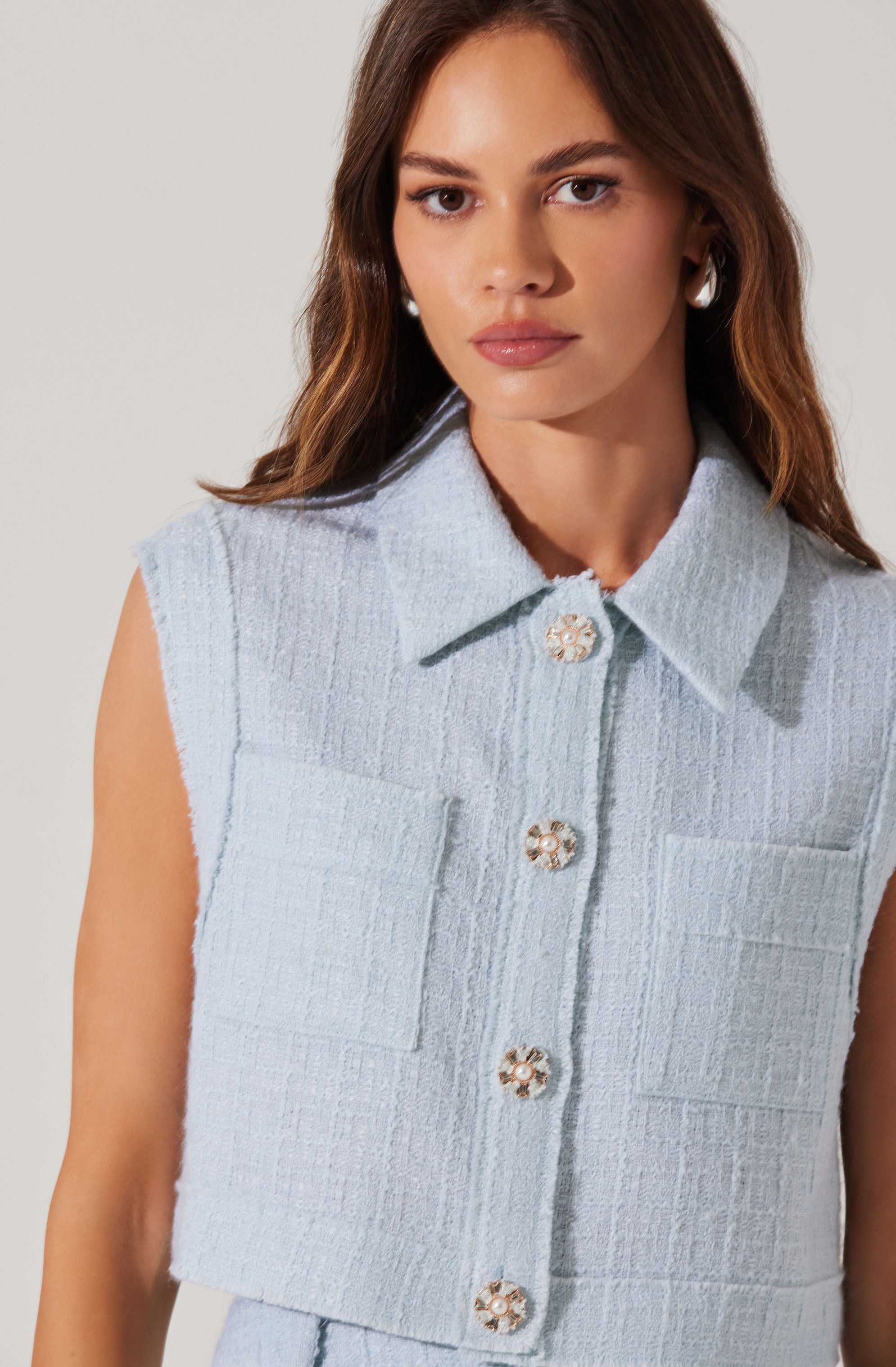 A woman wearing a sleeveless light blue cropped jacket with a textured fabric. The jacket features a classic collar and decorative gold buttons with floral designs down the front. She is also wearing large pearl earrings.
