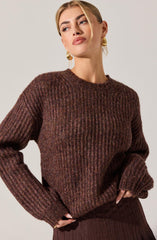 A woman is wearing a dark brown, ribbed knit sweater with a slightly loose fit. The sweater has a crew neckline and long sleeves that extend past her wrists. She is also wearing large, gold, hoop earrings.