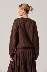 A woman is wearing a loose-fitting, long-sleeved sweater with a multicolored ribbed pattern in shades of brown, maroon, and hints of other earthy tones. The sweater has a round neckline and extends slightly below her waist. She pairs it with a high-waisted, pleated skirt in a matching dark brown color.
