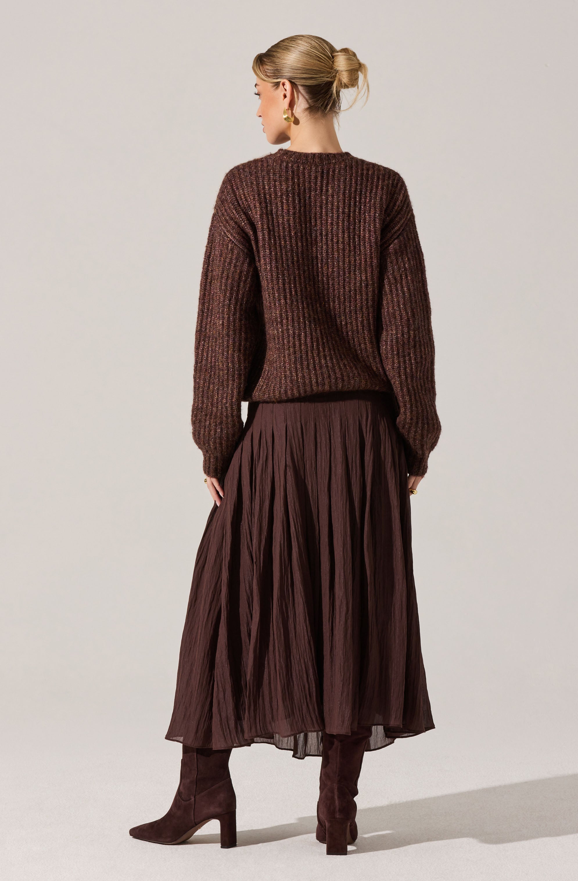 A woman is wearing a brown, chunky knit sweater with a ribbed texture, paired with a long, pleated, dark brown skirt. She has on brown suede high-heeled boots and gold earrings, with her blonde hair styled in a low bun.