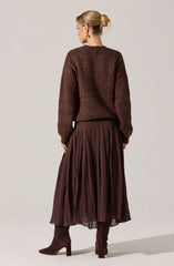 A woman is wearing a brown, chunky knit sweater with a ribbed texture, paired with a long, pleated, dark brown skirt. She has on brown suede high-heeled boots and gold earrings, with her blonde hair styled in a low bun.