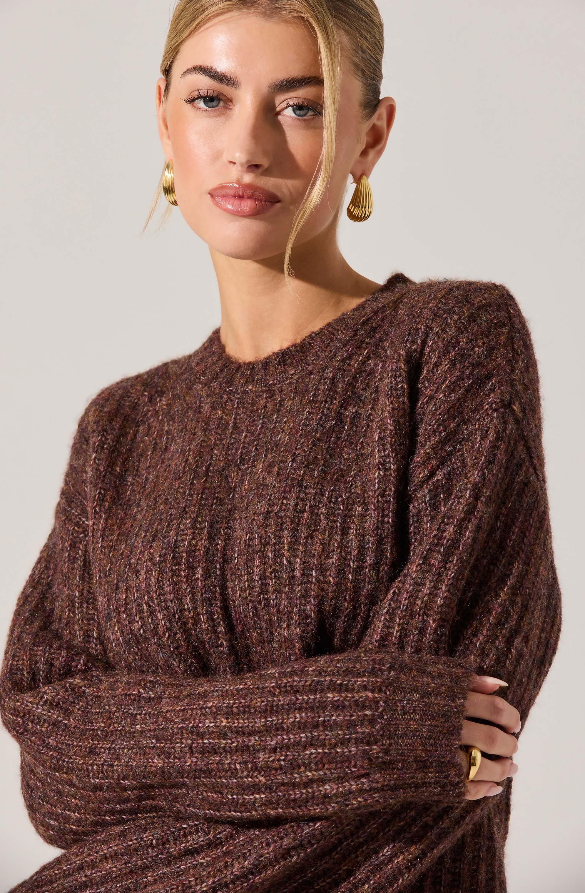 A woman is wearing a cozy, oversized sweater with a marled brown, purple, and pink color palette. The sweater has a ribbed, textured knit pattern and long sleeves. She is accessorized with large, gold, Egyptian-style hoop earrings and a gold ring on her right hand. Her hair is styled in a sleek, blonde updo, and her makeup is natural with a focus on her eyebrows and lips.