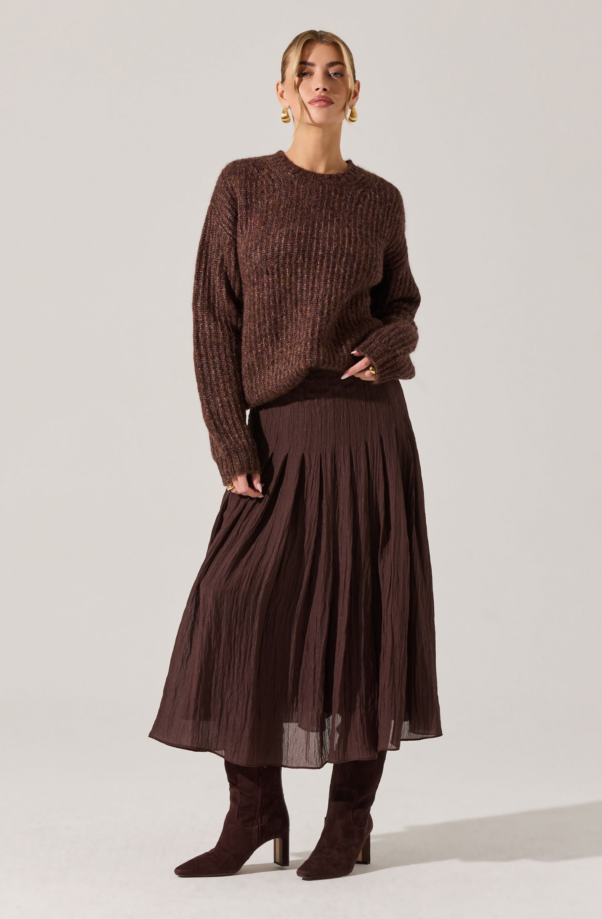 A woman is wearing a loose-fitting, textured, brown knitted sweater with a round neckline and long sleeves. She pairs it with a dark brown, pleated, midi-length skirt made of a lightweight, semi-sheer fabric. She completes the outfit with matching dark brown suede high-heeled boots and gold hoop earrings.