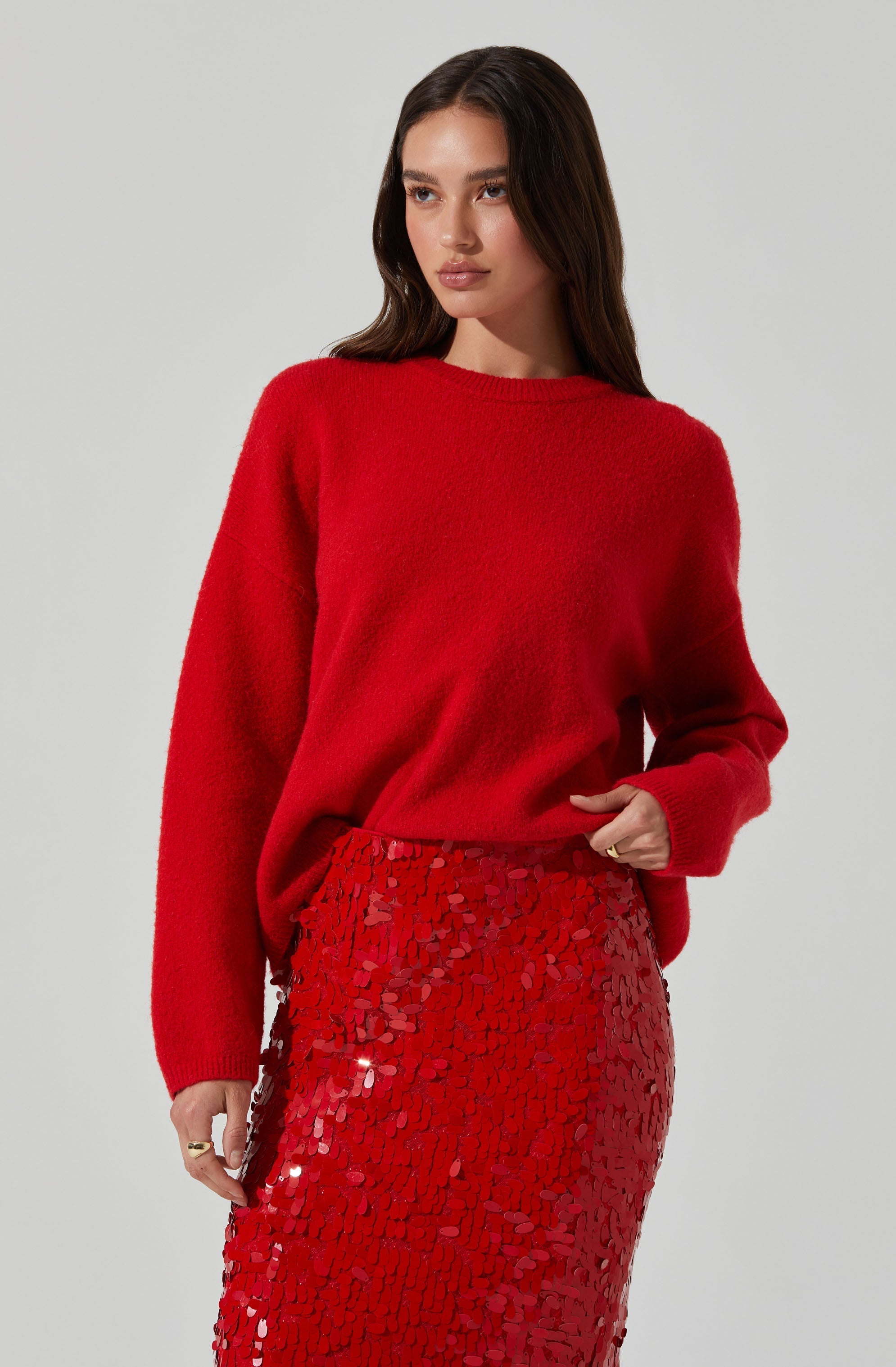 A woman is wearing a bright red, long-sleeved, fuzzy sweater with a round neckline, paired with a high-waisted, fitted skirt covered in shiny, red sequin embellishments.
