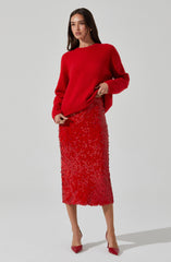 A woman is wearing a bright red, long-sleeved, oversized sweater paired with a red textured midi skirt that has a floral or abstract pattern. She completes her outfit with red pointed-toe heels.