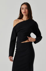 The woman is wearing an off-the-shoulder black top with long sleeves, paired with a high-waisted black skirt. She accessorizes with a chunky gold chain necklace and a ring on her right hand.