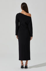 The outfit is a black, long-sleeved dress with an off-the-shoulder design on the left side, exposing the shoulder and collarbone. The dress is fitted at the waist and extends down to the ankles. The wearer is also wearing black high-heeled shoes with an ankle strap.