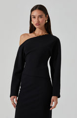 A woman wearing a black, off-the-shoulder dress with long sleeves. The dress has a smooth, fitted silhouette. She accessorizes with gold hoop earrings, a chunky gold necklace, and rings on her fingers.