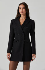 The woman is wearing a tailored black blazer dress with a double-breasted design, featuring six buttons on the front. The blazer has a notched lapel collar, structured shoulders, and long sleeves. The dress length is above the knees, revealing sheer black tights.