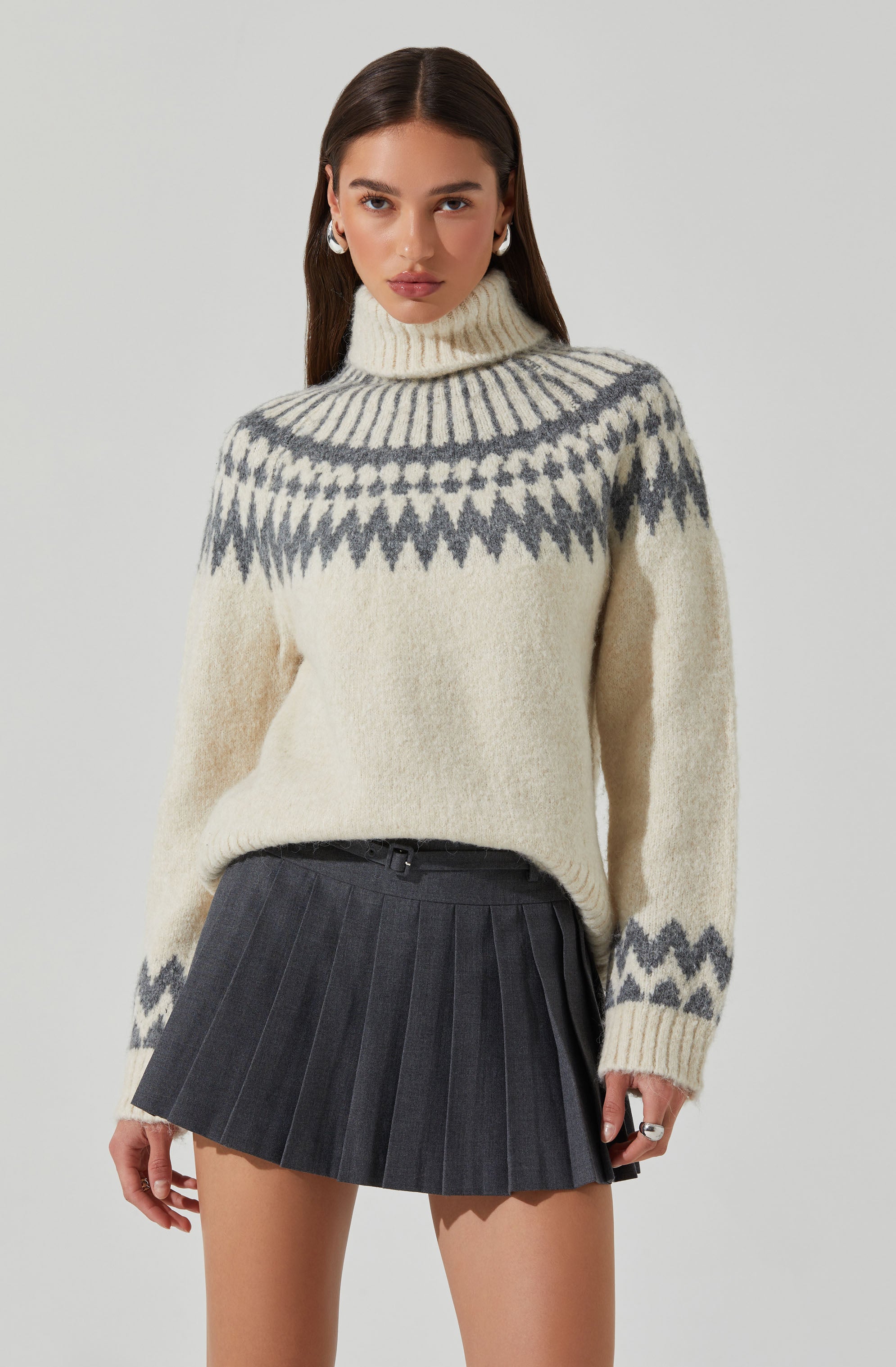 A woman is wearing a cream-colored, chunky knit turtleneck sweater with a gray geometric pattern around the yoke, cuffs, and hem. She pairs it with a high-waisted, black pleated mini skirt featuring thin vertical stripes.