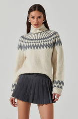 A woman is wearing a cream-colored, chunky knit turtleneck sweater with a gray geometric pattern around the yoke, cuffs, and hem. She pairs it with a high-waisted, black pleated mini skirt featuring thin vertical stripes.
