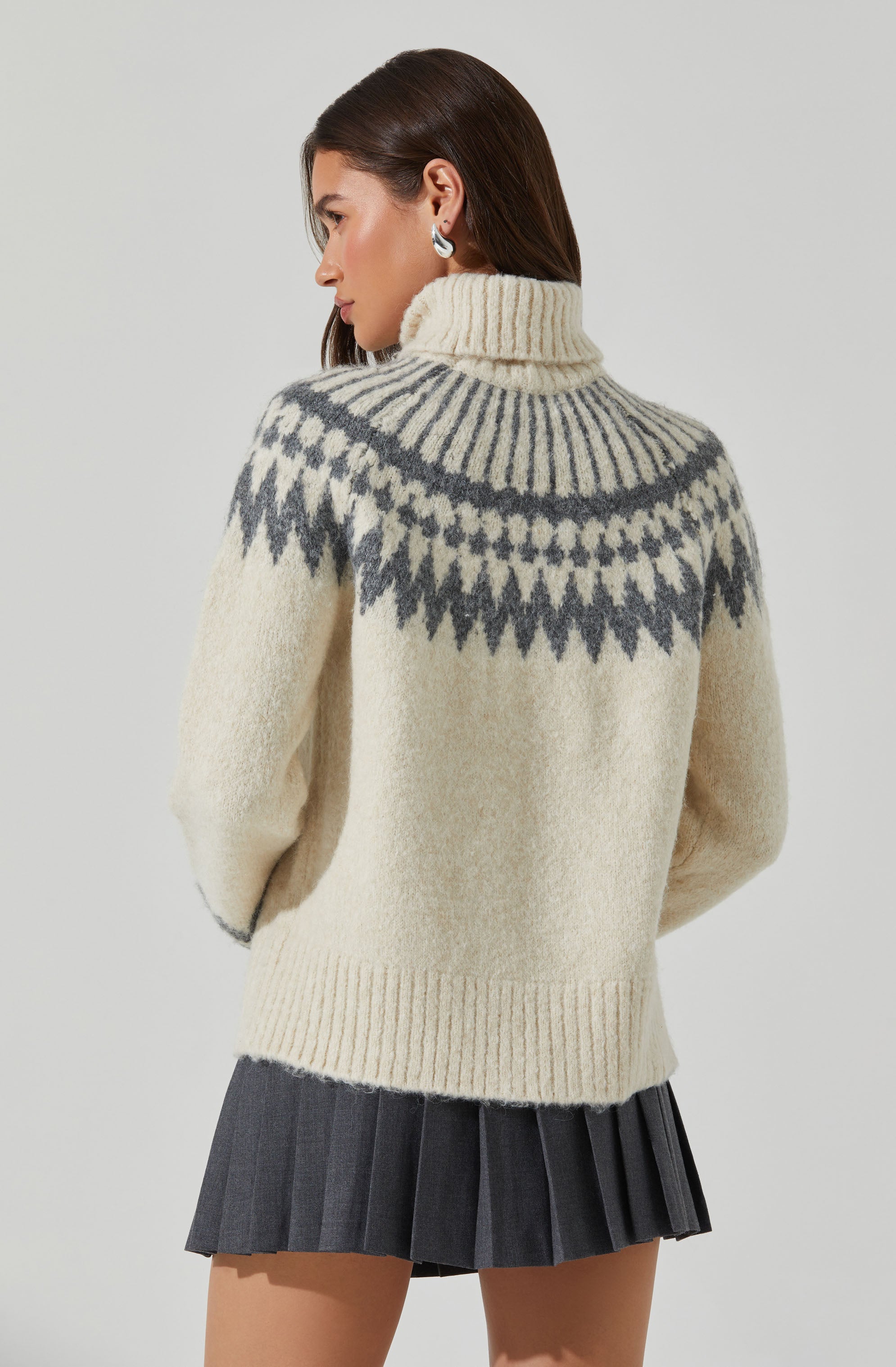 A woman is wearing a cozy, oversized turtleneck sweater with a cream base color and a dark gray geometric pattern around the shoulders and neckline. The sweater has a ribbed hem and cuffs. She pairs the sweater with a dark gray pleated skirt.