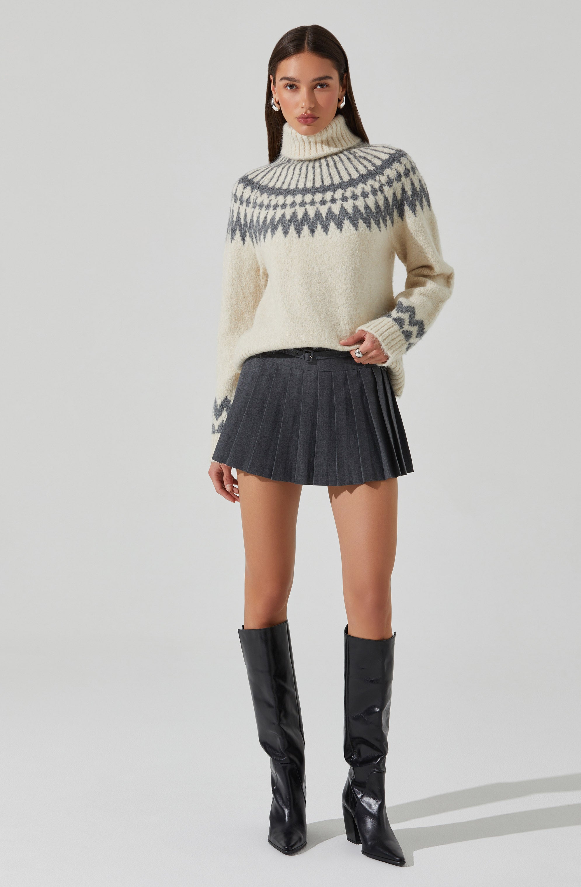 A woman is wearing a cream-colored knit turtleneck sweater with a gray geometric pattern around the shoulders and upper chest. She has paired it with a high-waisted, dark gray pleated mini skirt and black knee-high boots with pointed toes and high heels.
