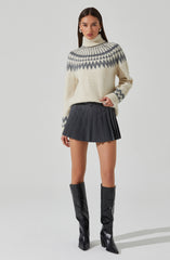 A woman is wearing a cream-colored knit turtleneck sweater with a gray geometric pattern around the shoulders and upper chest. She has paired it with a high-waisted, dark gray pleated mini skirt and black knee-high boots with pointed toes and high heels.
