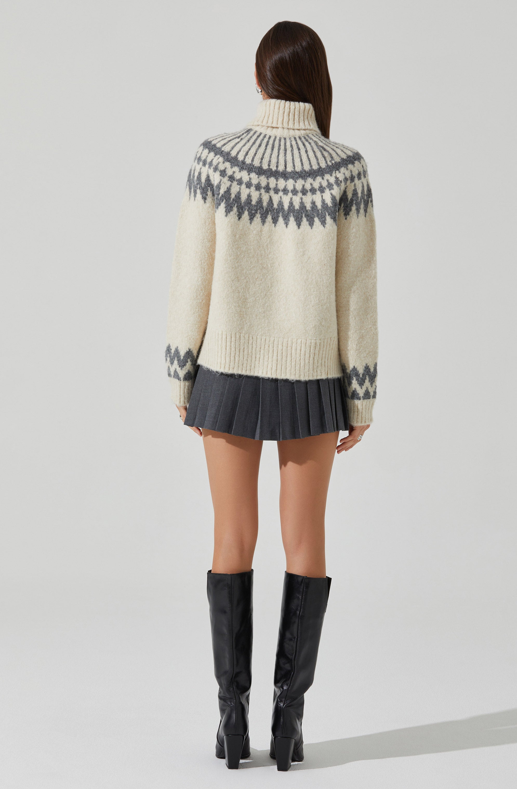 A person is wearing a cream-colored, chunky knit turtleneck sweater with a gray geometric pattern across the shoulders and sleeves. The sweater has a relaxed fit, with the pattern featuring zigzags and lines. They are also wearing a dark gray pleated mini skirt and tall black leather heeled boots.