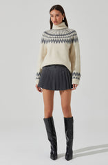 A woman is wearing an oversized, cream-colored turtleneck sweater with a gray and white geometric pattern around the yoke and sleeves. She pairs it with a high-waisted, dark gray pleated mini skirt. She completes the outfit with black, knee-high boots with a shiny finish.