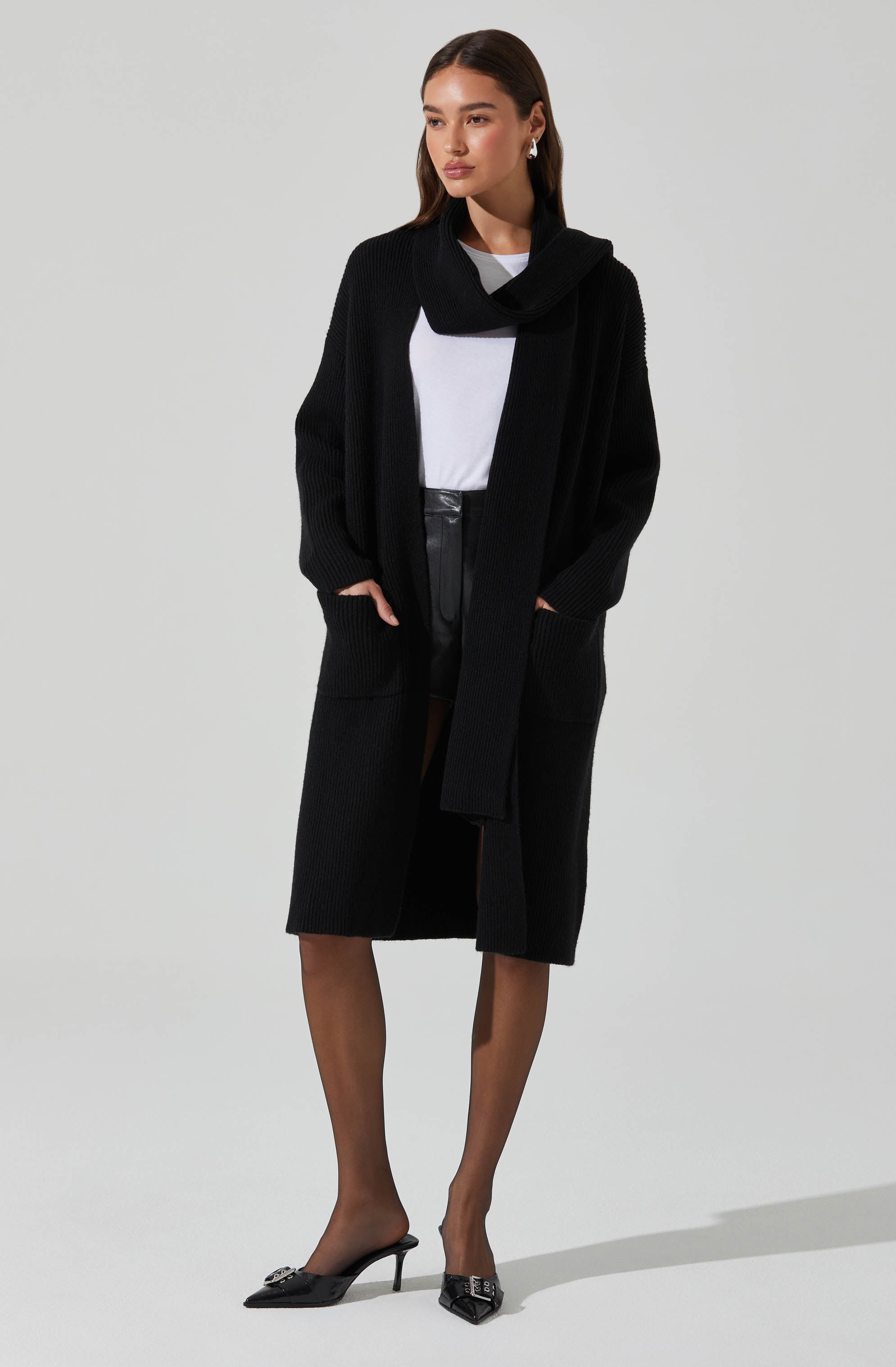 A woman is wearing a long black cardigan with large front pockets, a white top underneath, and black leather shorts. She has sheer black tights and black pointed-toe high heels with decorative embellishments. Her hair is styled straight, and she is wearing earrings.