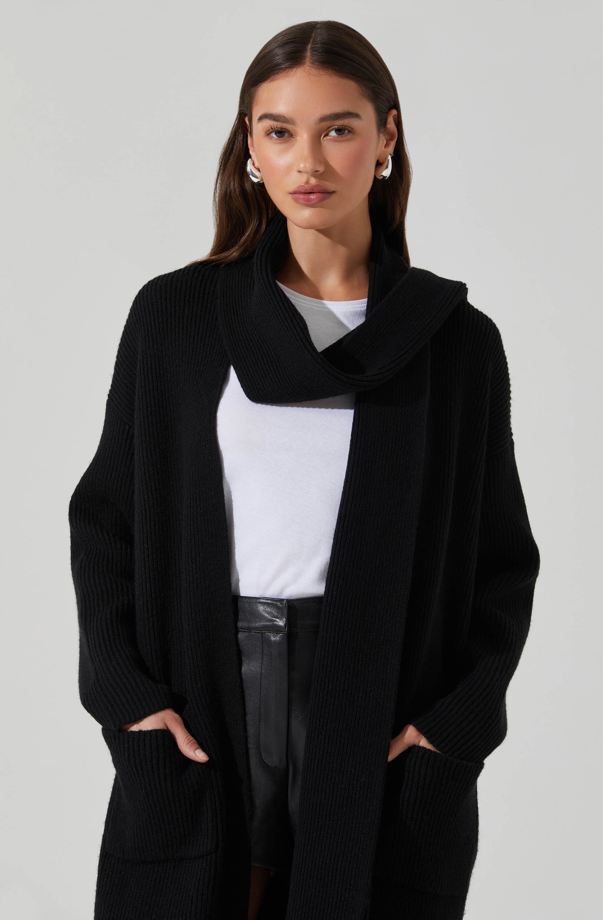 A woman is wearing a long, oversized black knit cardigan with large front pockets. She has on a white t-shirt underneath and high-waisted black shorts made of a shiny material. The cardigan features a wide collar that drapes around her neck.