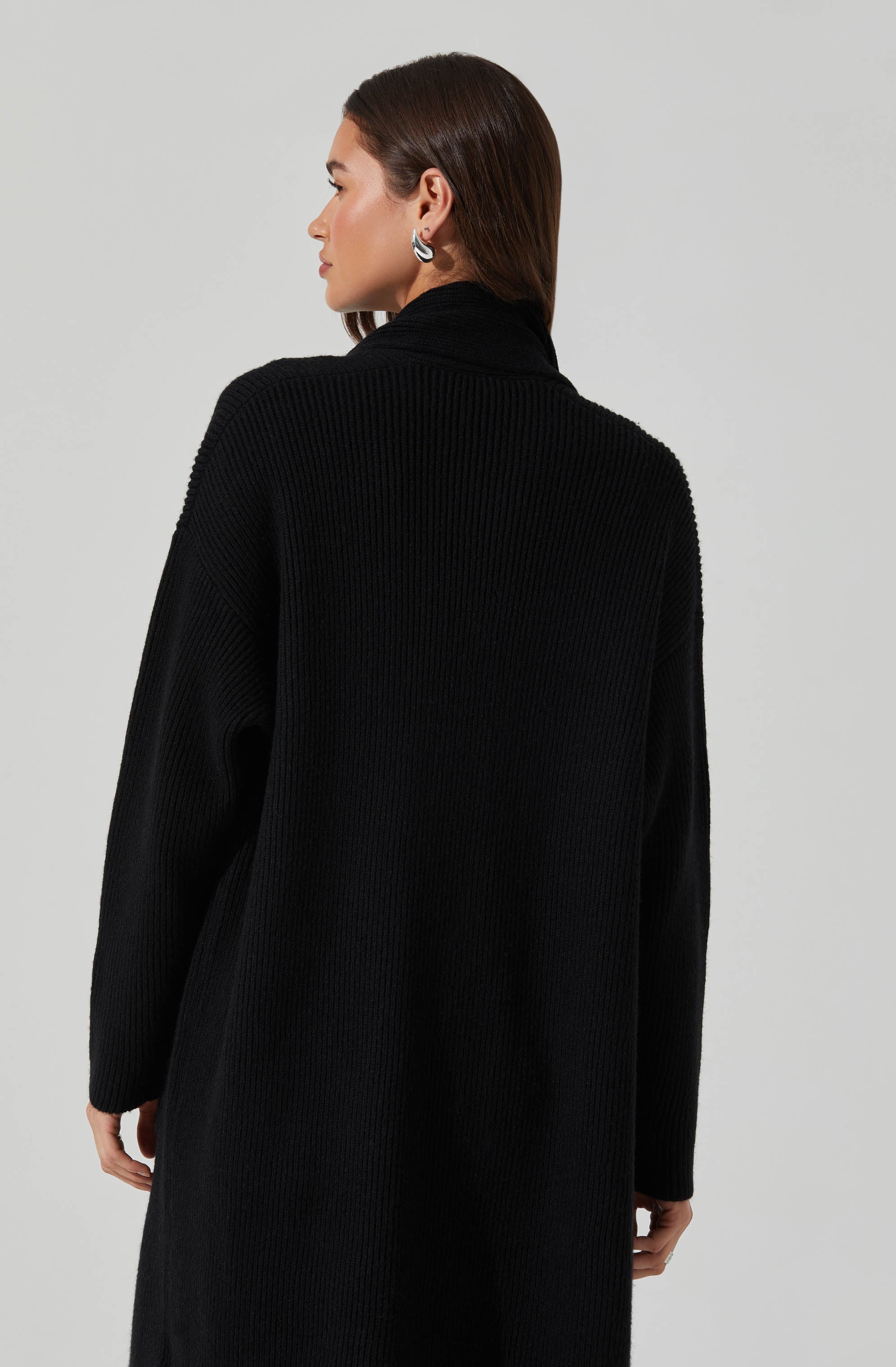 A woman is wearing a black, ribbed knit coat with a high collar. The coat appears to be oversized and has long sleeves. She has dark brown hair and is accessorizing with silver hoop earrings.