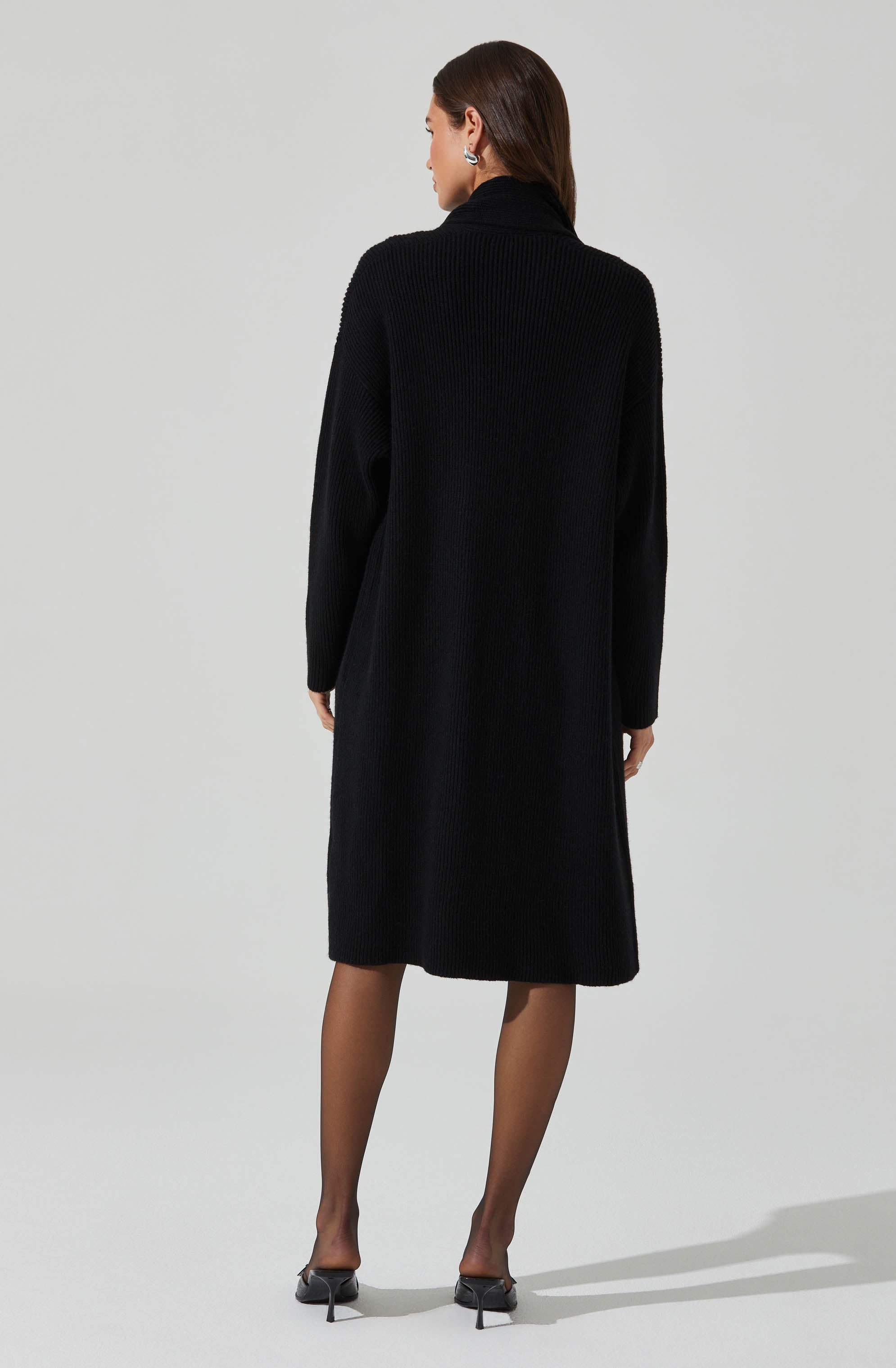 A woman is wearing a long, black, ribbed knit coat with a high collar. She has on sheer black stockings and black high-heeled shoes. She is also wearing large, silver, circular earrings.