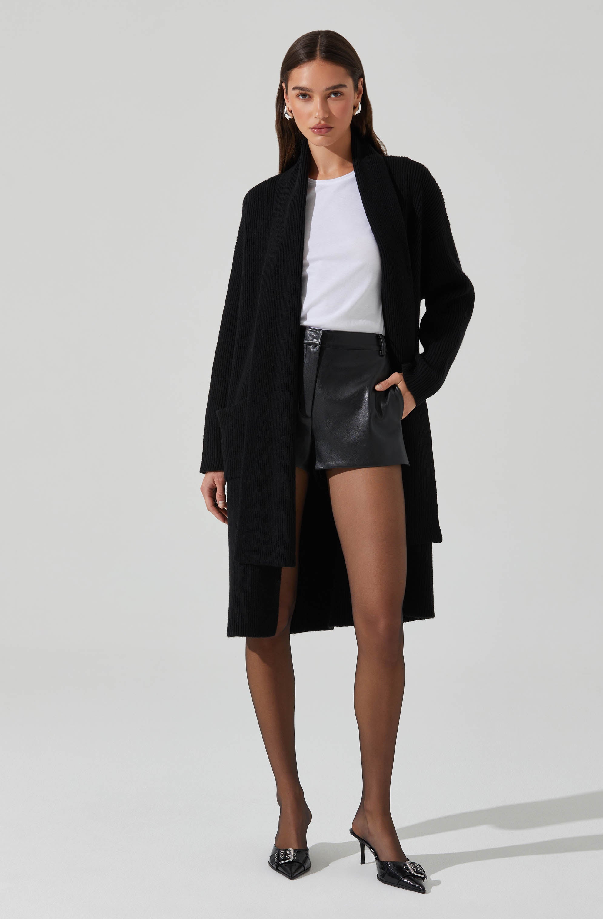 A woman is wearing a long black knit cardigan with large pockets, a white T-shirt, black leather shorts, sheer black tights, and black high-heeled shoes with pointed toes and embellishments.