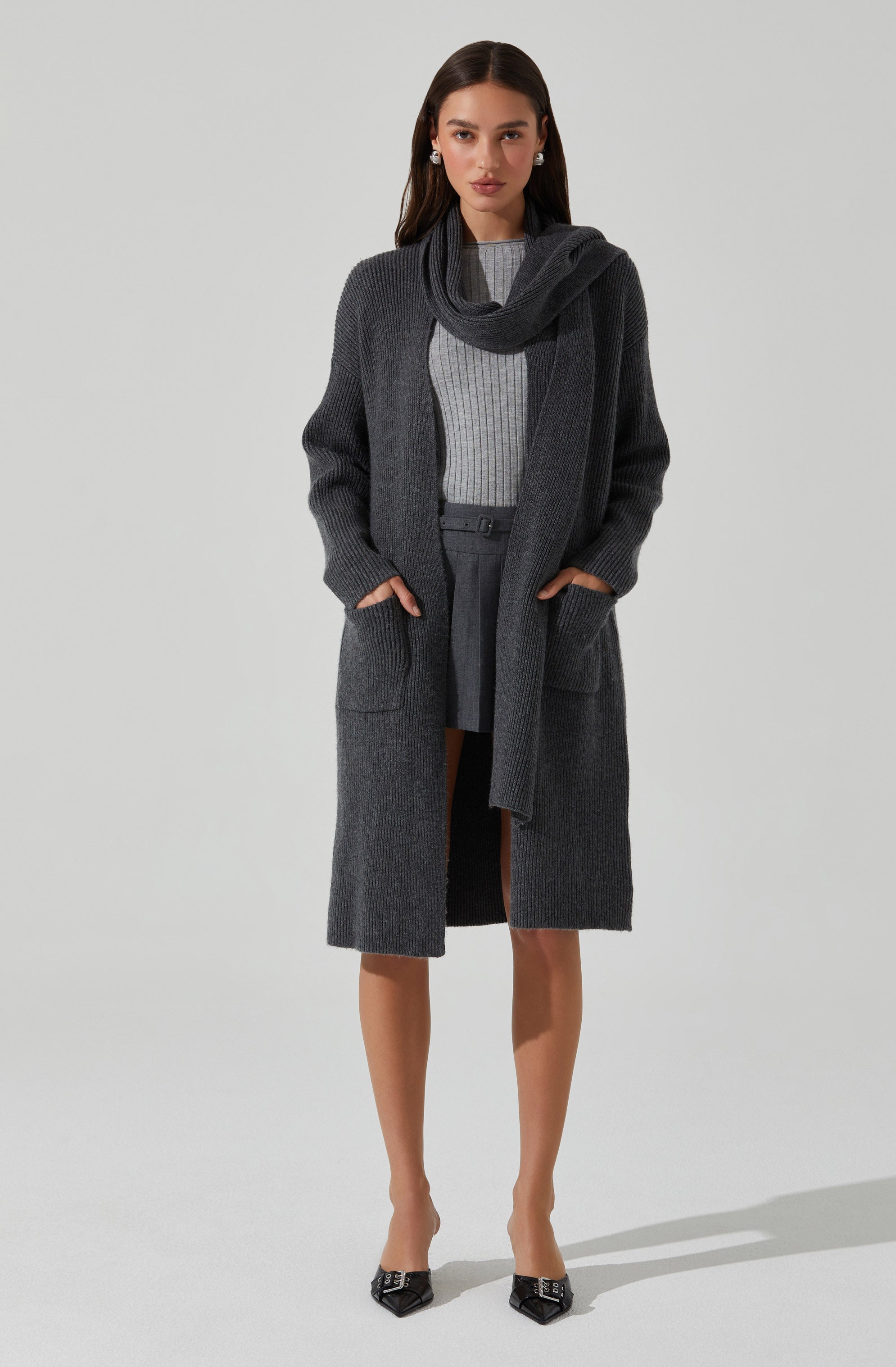 A woman is wearing a dark gray, long, knit cardigan with large front pockets, layered over a light gray, ribbed sweater. She has a matching dark gray scarf draped around her neck. She is also wearing dark gray high-waisted pants and black pointed-toe heels with metallic embellishments.