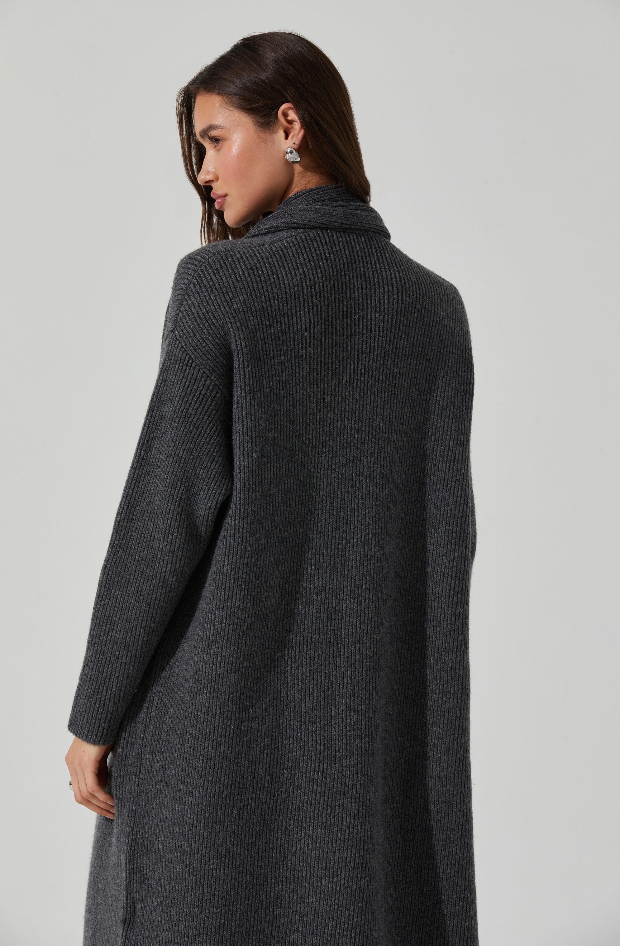 A woman is wearing a long, dark gray, ribbed knit sweater with a high collar. The sweater appears cozy and is made of a thick, textured fabric. She has dark, straight hair and is accessorized with earrings.