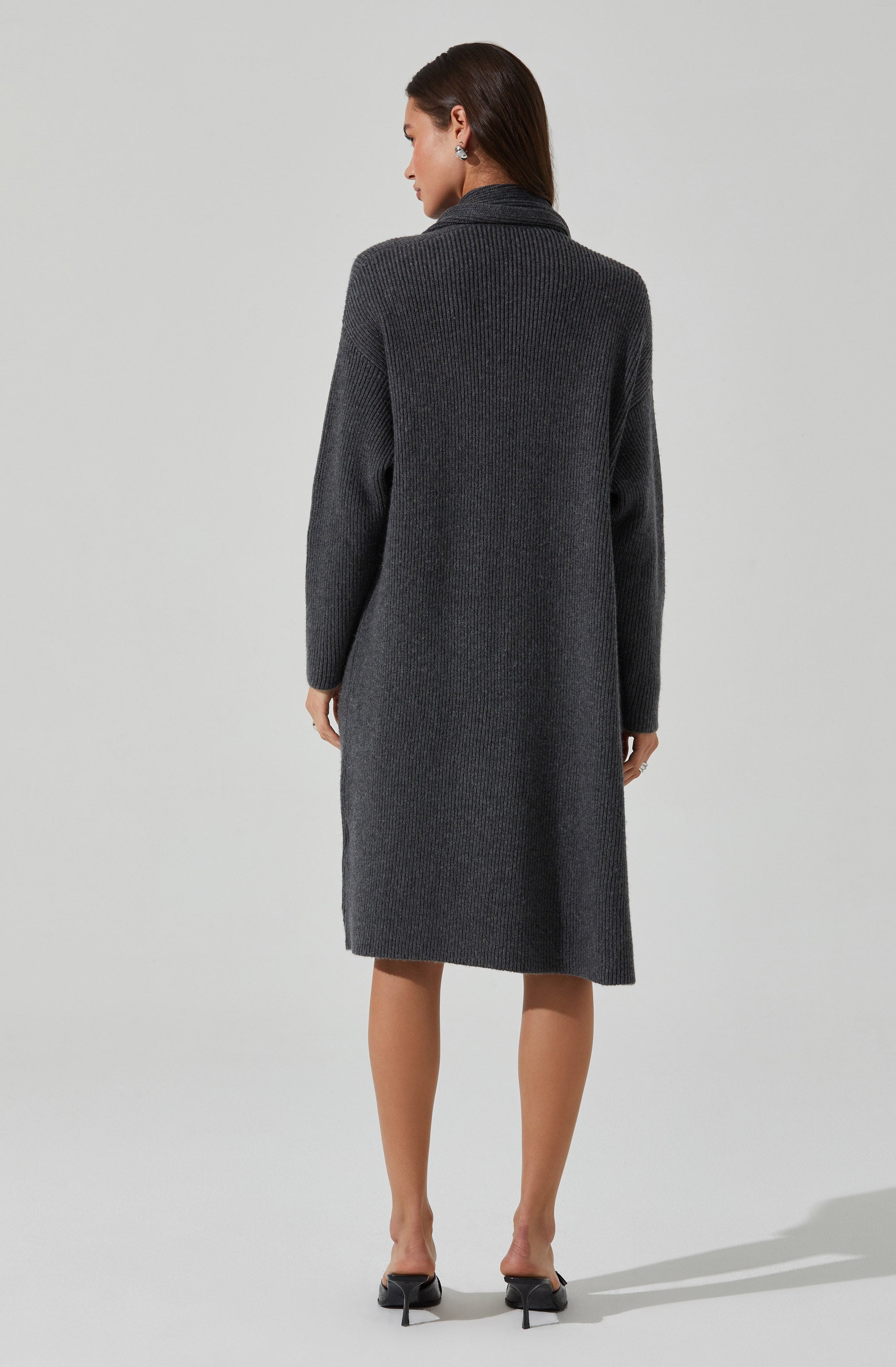 A woman is wearing a long, dark gray, knitted cardigan or coat that reaches below her knees. The garment has a textured, ribbed pattern and a relaxed fit with long sleeves. She is also wearing black pointed-toe high heels.