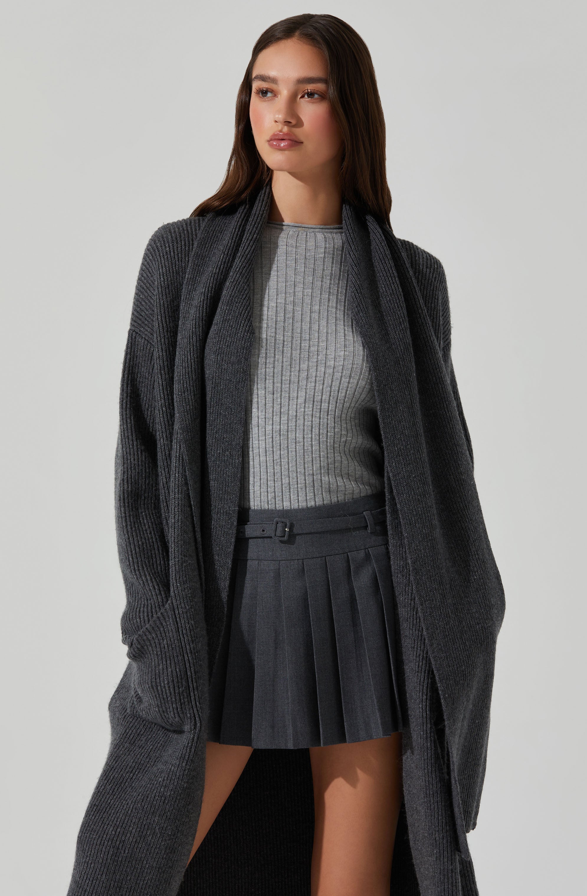 A woman is wearing a long, dark gray, ribbed knit cardigan over a light gray, ribbed long-sleeve top. She is also wearing a high-waisted, dark gray pleated skirt with a belt detail.