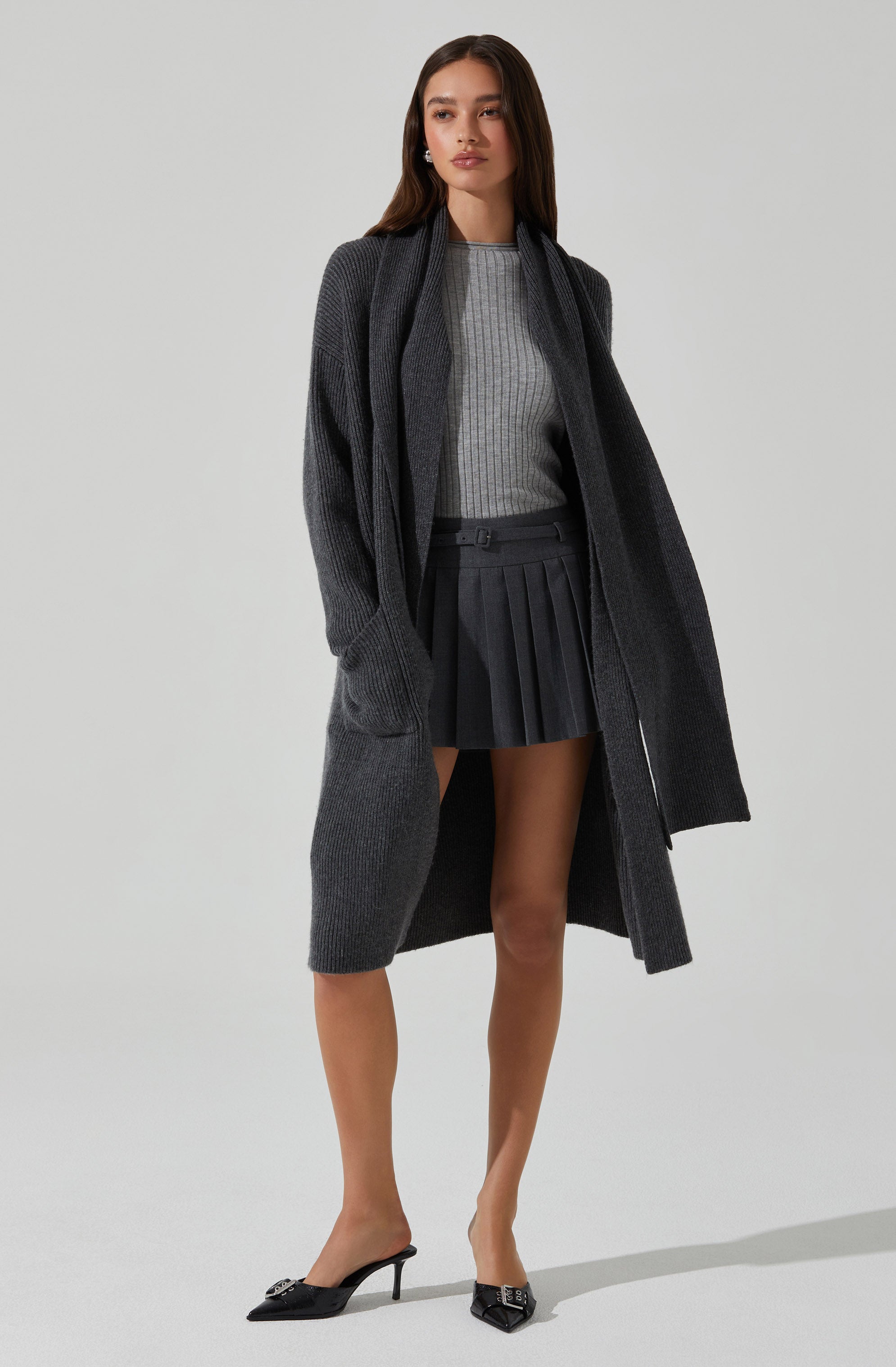 A woman wearing a long, dark gray knitted cardigan over a light gray, ribbed knit sweater. She has on a high-waisted, pleated black mini skirt and black pointed-toe high heels.