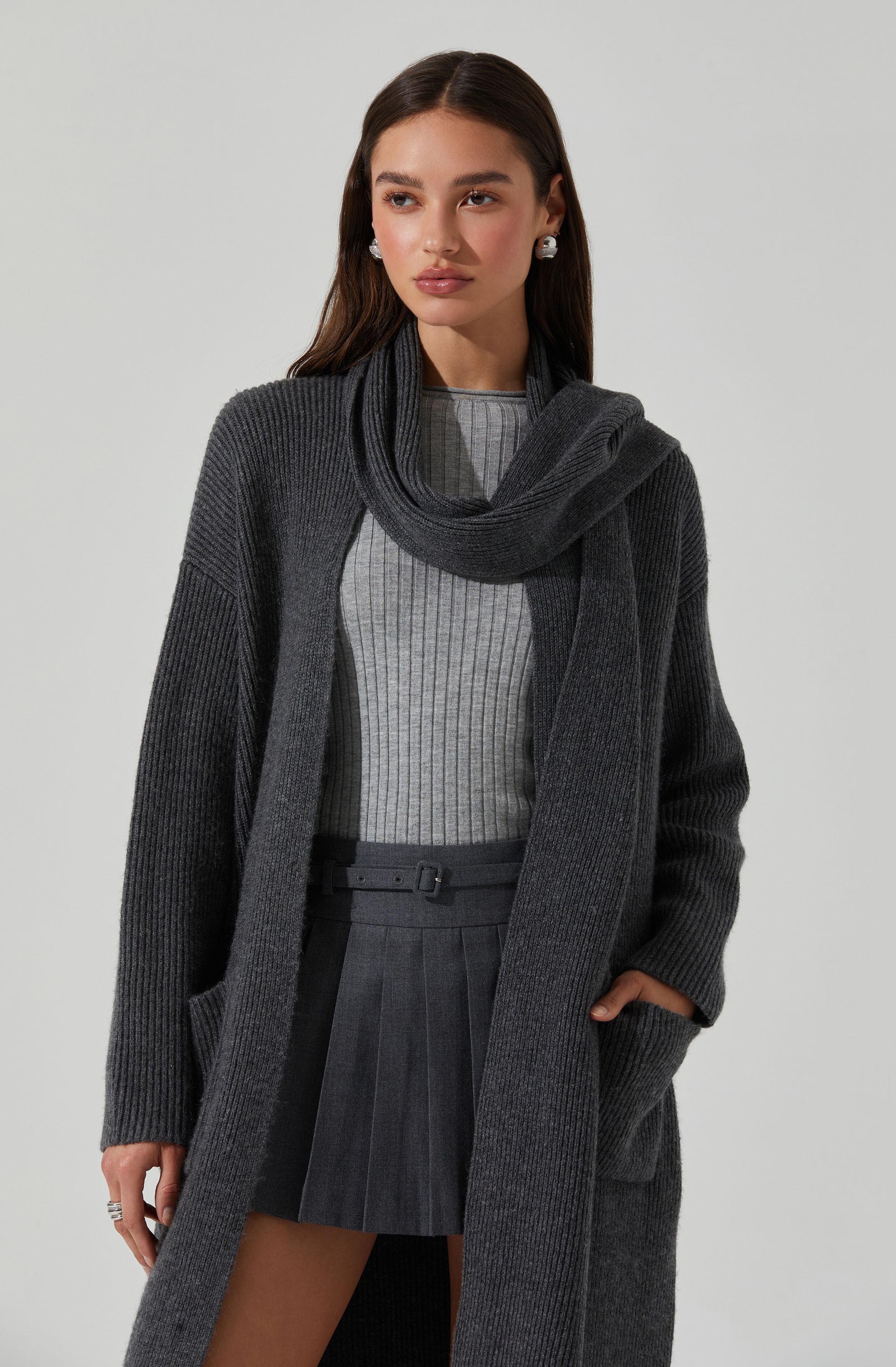 A woman is wearing a dark gray, ribbed-knit long cardigan with large pockets. She has a matching gray scarf draped around her shoulders and a light gray, vertically striped top underneath. She is also wearing a high-waisted, pleated dark gray skirt with a belt.