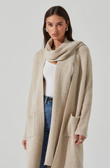 A woman is wearing a long, beige, ribbed knit cardigan with large front pockets. She has a matching beige scarf draped around her shoulders, a white top underneath, and blue jeans.