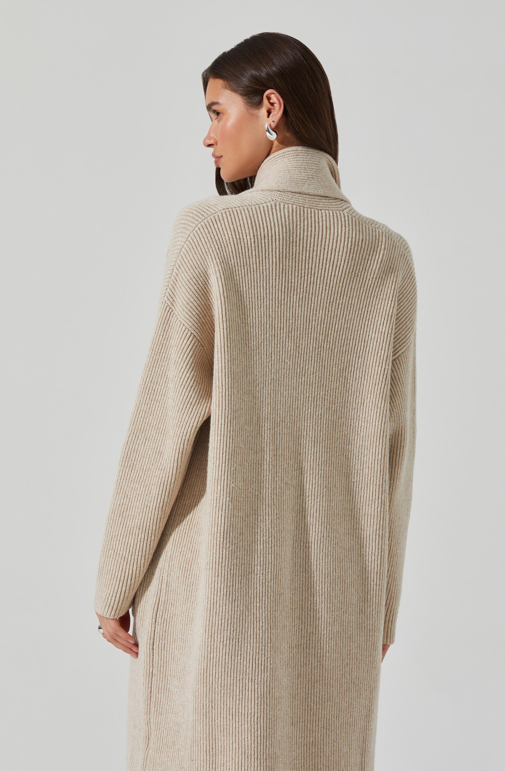 A woman is wearing a long, beige, ribbed knit coat with a high collar. The coat appears cozy and oversized, with relaxed fit and dropped shoulders. She has dark, straight hair and is accessorized with silver hoop earrings.