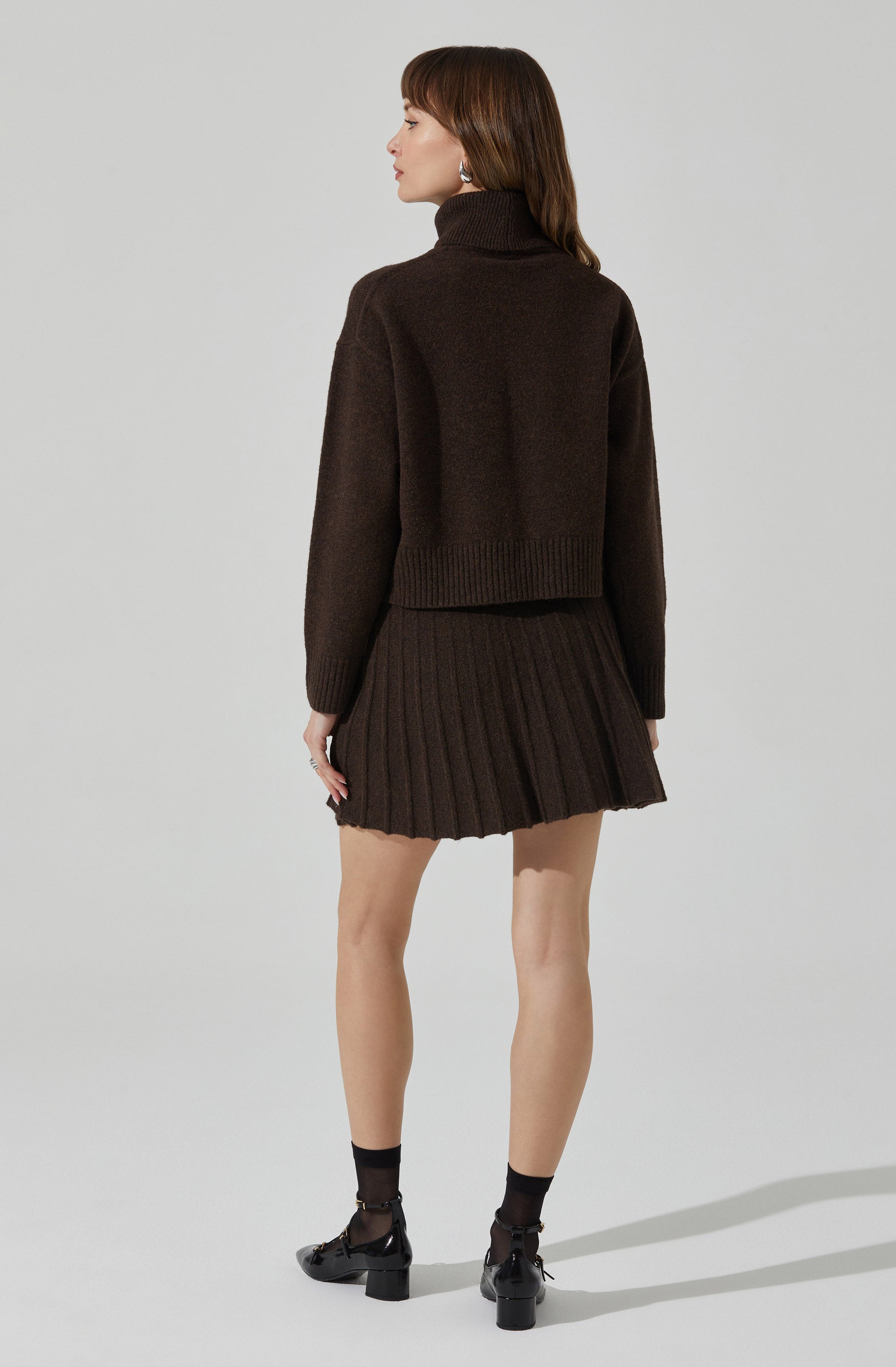 A woman is wearing a dark brown, chunky knit turtleneck sweater with a loose fit and ribbed hem. She pairs it with a matching dark brown pleated mini skirt. She is also wearing black ankle socks and black heeled shoes with ankle straps.