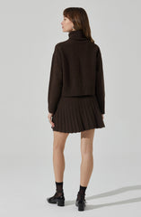 A woman is wearing a dark brown, chunky knit turtleneck sweater with a loose fit and ribbed hem. She pairs it with a matching dark brown pleated mini skirt. She is also wearing black ankle socks and black heeled shoes with ankle straps.