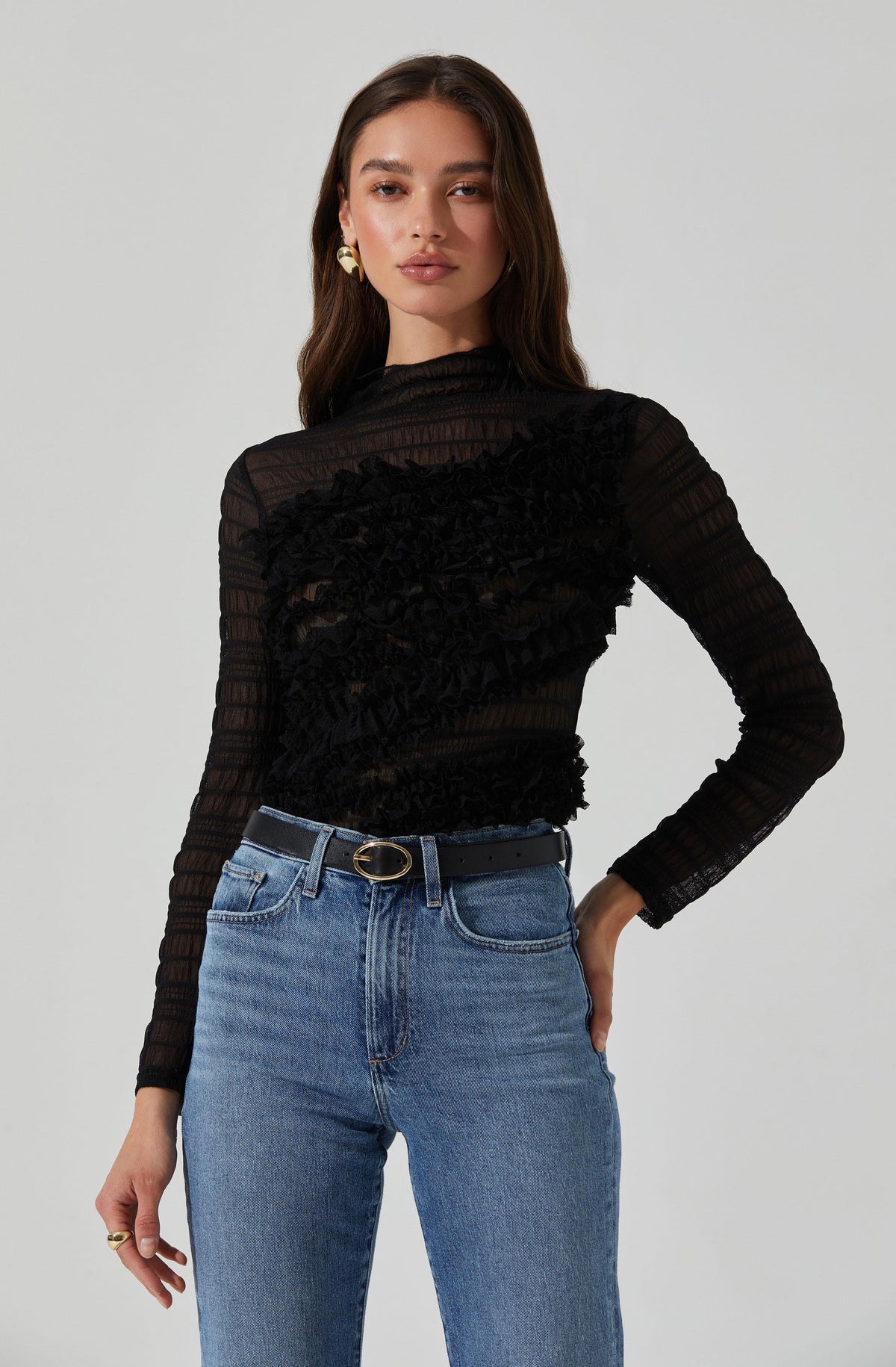 A woman is wearing a long-sleeved, semi-sheer black top with ruffled details across the front and vertical stripes. She pairs the top with high-waisted, light blue jeans and a black belt with a gold buckle. She has on gold earrings and a ring.