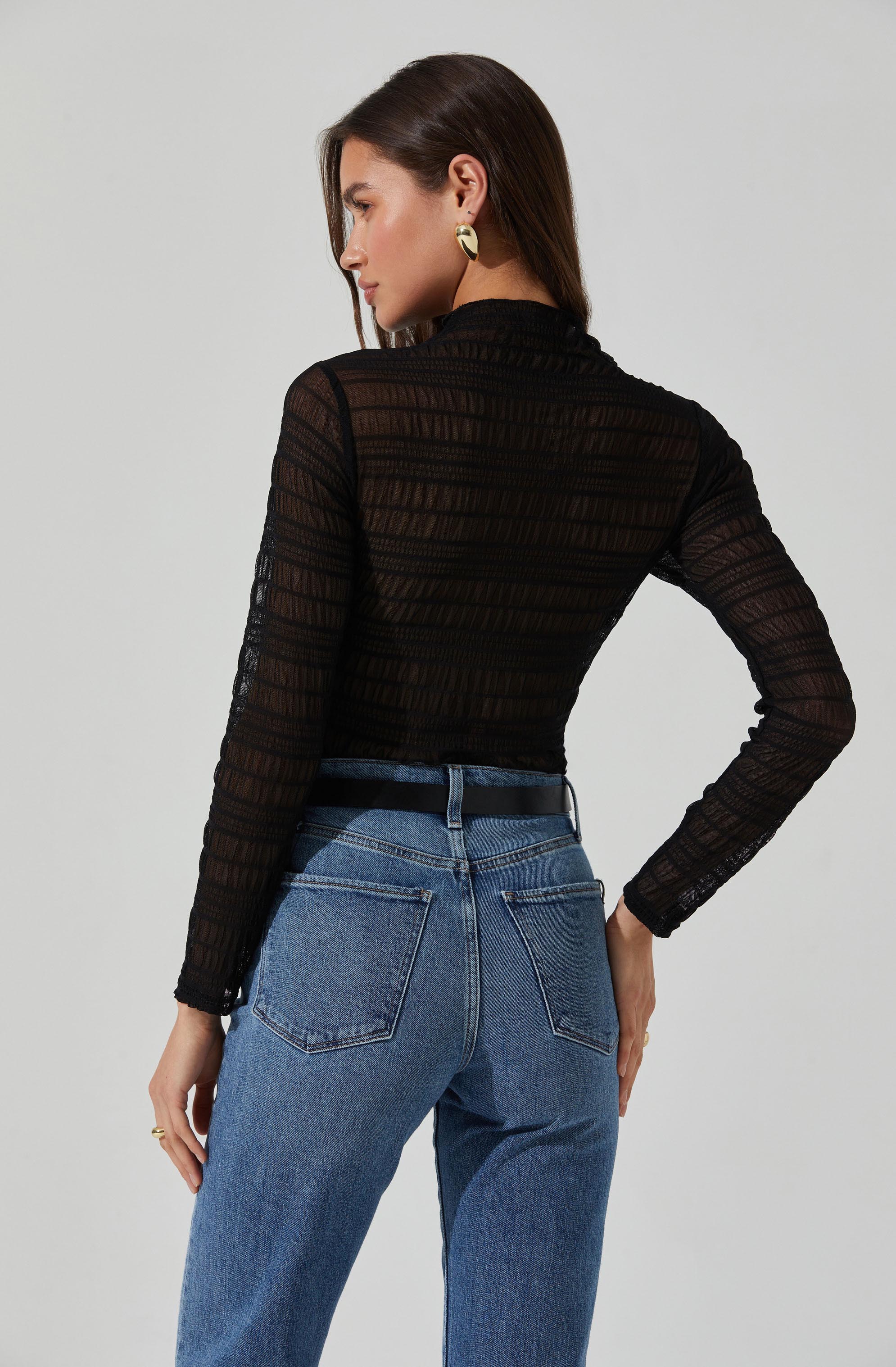 A woman is wearing a long-sleeve black sheer top with horizontal striped patterns and a high collar. She pairs the top with high-waisted blue jeans that have prominent back pockets and a black belt. She has medium-length dark hair and is accessorized with large gold earrings.